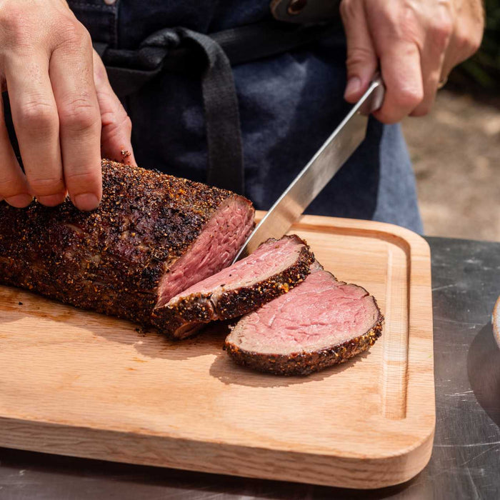 One hand holding the top of a cooked slow-roasted filet mignon, the other hand slicing the steak with a large knife on a wooden cutting board on top of an Ooni Modular Table.