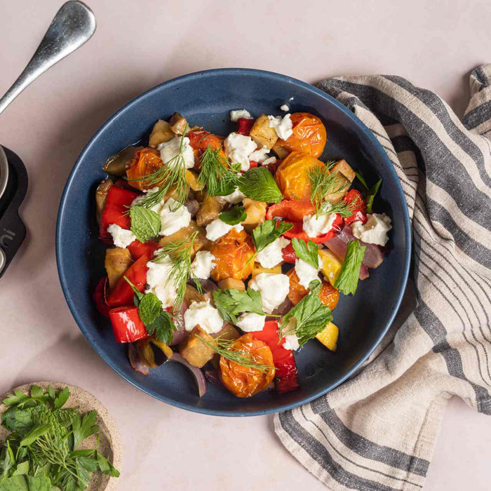 Slow-roasted squash, aubergine, onions, cherry tomatoes and sweet peppers in an Ooni Roasting pan next to a bowl of slow-roasted vegetables with feta and fresh herbs on a table.