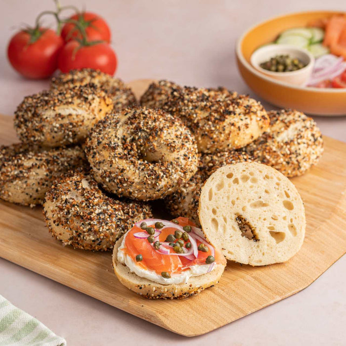 A stack of unsliced everything bagels and one bagel sliced and spread with cream cheese, lox, onions and capers on an Ooni Bamboo Pizza Peel