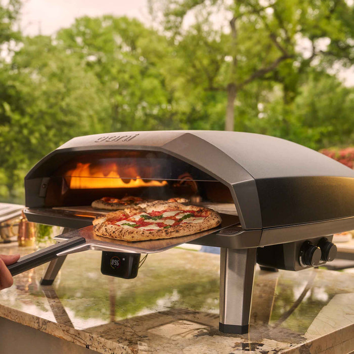 Ooni Koda 2 Max outdoor pizza oven cooking two pizzas, one already in the oven and another being inserted on a pizza peel in a backyard. | Cliquez sur cette image pour ouvrir la fenêtre modale de produits. La fenêtre modale de produits permet de zoomer sur les images.