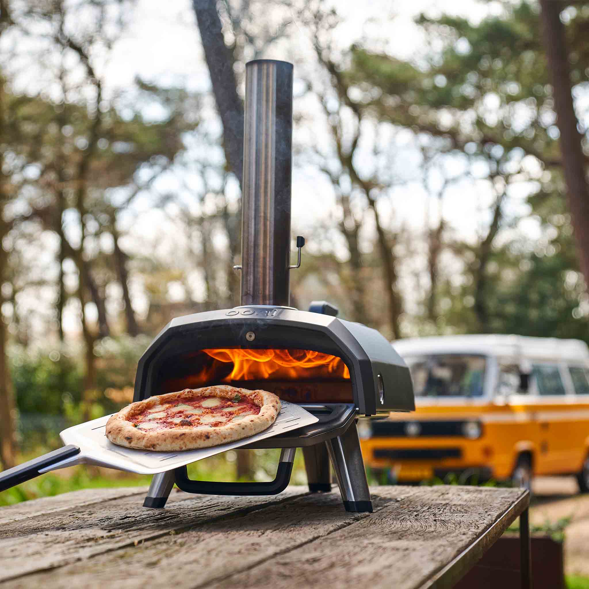 Ooni Karu 2 multi-fuel pizza oven  on a park bench. pizza in shot as well as camper van in background. 